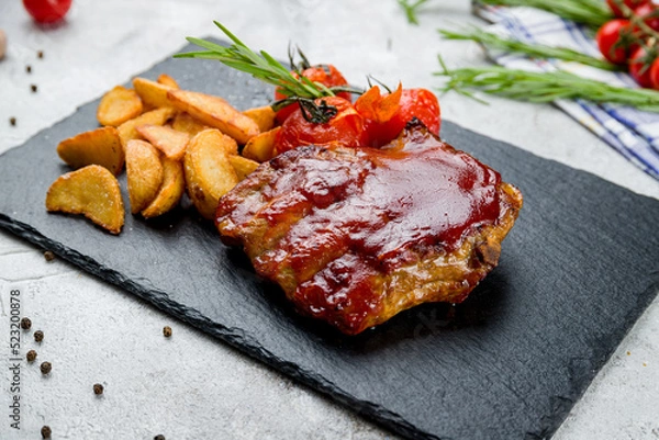 Obraz Barbecue pork ribs with potato wedges and tomatoes on black stone plate on grey table macro close up
