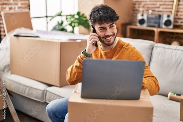 Fototapeta Young hispanic man using laptop talking on smartphone at new home