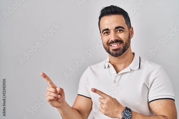 Fototapeta Young hispanic man with beard wearing casual clothes over white background smiling and looking at the camera pointing with two hands and fingers to the side.