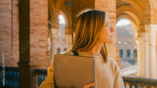 Fototapeta Portrait shot of beautiful young woman walk in sunny corridor of campus building. Female student or professor hold papers and laptop. Education concept. Study abroad. Cinematic real candid portrait