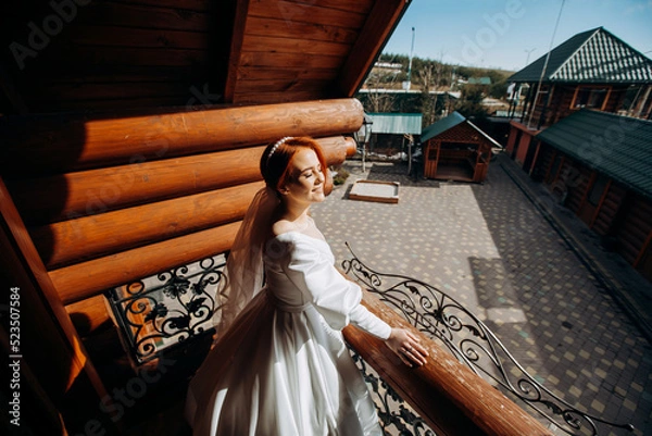 Fototapeta Luxury red-haired bride in a satin white dress is posing on the balcony on her wedding day.