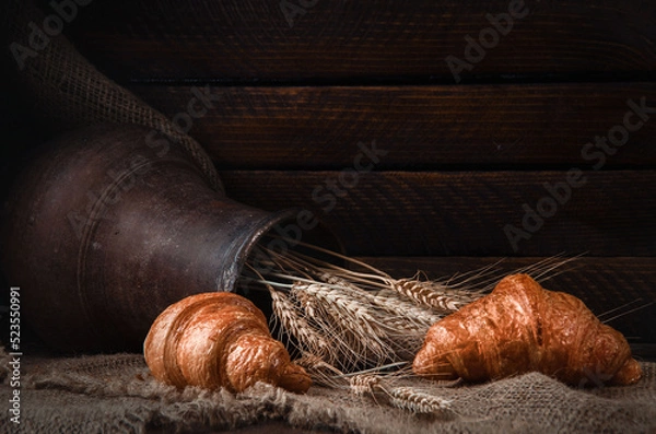 Fototapeta still life earthenware jug with wheat ears, bread, croissants on a drapery