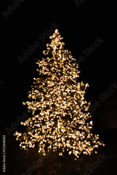 Fototapeta Silhouette of blurry Christmas tree illuminated and decorated with golden fairy lights in dark night creating beautiful bokeh effect with glowing circles or shiny dots, abstract image for holiday card
