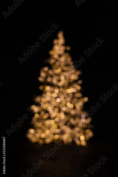 Fototapeta Silhouette of blurry Christmas tree illuminated and decorated with golden fairy lights in dark night creating beautiful bokeh effect with glowing circles or shiny dots, abstract image for holiday card