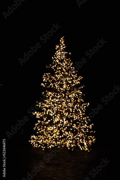 Fototapeta Silhouette of blurry Christmas tree illuminated and decorated with golden fairy lights in dark night creating beautiful bokeh effect with glowing circles or shiny dots, abstract image for holiday card