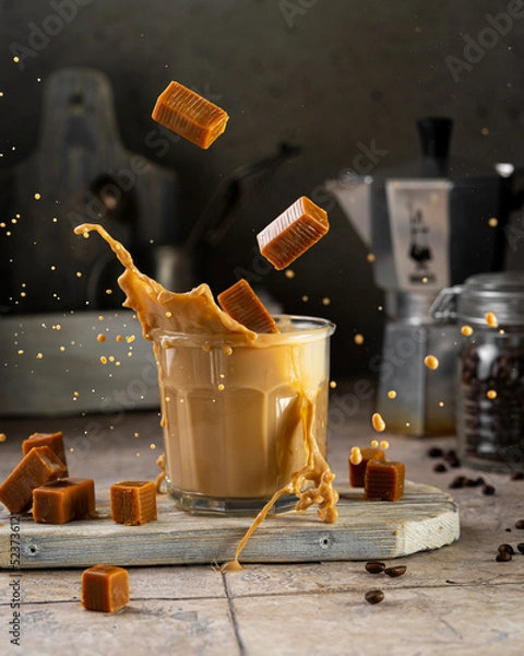 Fototapeta Coffee with milk (caramel) splash in glass with toffee bites, flying food concept. Sweet drink dessert on wooden board, beautiful studio image with copy space.