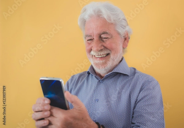 Obraz Portrait of beautiful smiling senior bearded man isolated on yellow background while using phone in video call