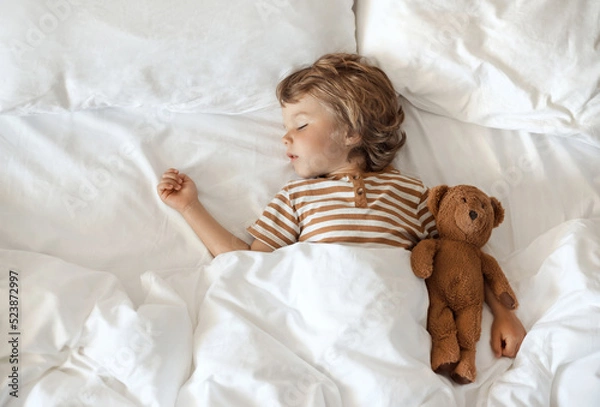 Fototapeta Top view of a cute little baby hugging his toy teddy bear and sweet sleeping under a blanket on a soft comfortable pillow, white clean bedding in cozy bed at home