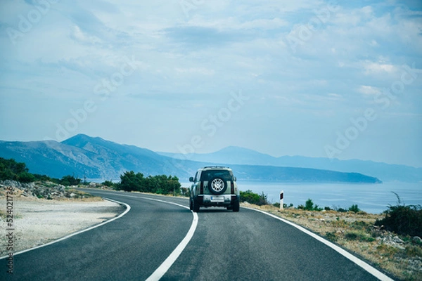 Fototapeta Rear view of SUV car driving on highway along the Adriatic sea with beautiful mountain view and blue clouds