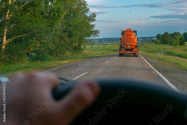 Fototapeta view of a fuel tanker with an orange tank carrying flammable cargo on a country road against the background of a driver who is driving behind a truck