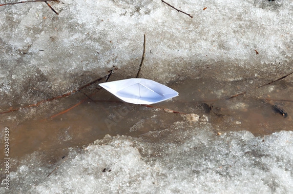 Fototapeta Top view of white paper boat sails along a spring stream among the snowy shores