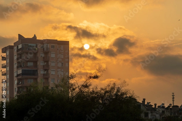 Obraz Orange sky at sunset, a high-rise building and the sun nearby, Clouds and trees.