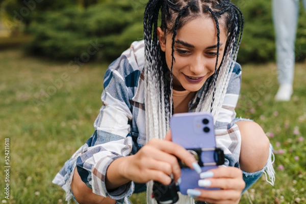 Obraz The girl with the afro pigtails arranges the phone in order to film herself in a tik tok video. Active life on the Internet. Followers drop.