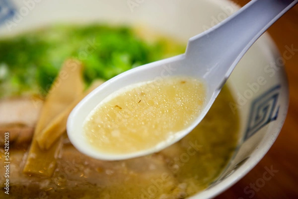 Obraz ラーメン店で提供される、背脂が浮いたコク深い黄金色の塩ラーメンスープ。レンゲですくい上げたシズル感あふれるアップ写真