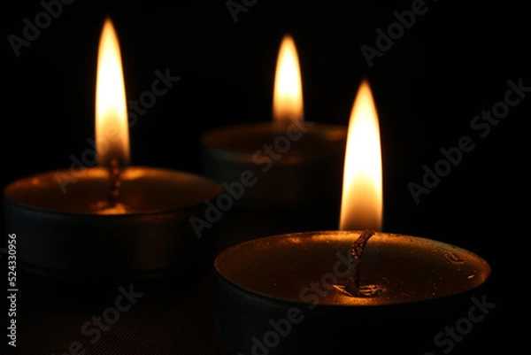Fototapeta Three burning candles close-up on a black background with copy space. Blurred background