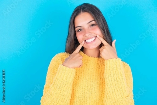 Fototapeta Happy beautiful brunette woman wearing yellow sweater over blue background with toothy smile, keeps index fingers near mouth, fingers pointing and forcing cheerful smile
