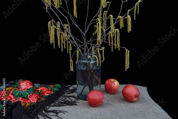 Fototapeta Retro still life with vintage shawl and a bouquet of spring birch branches with buds  in vase and red apples on  table  black background, concept of old rustic interiors, winter, arrival of Carnival