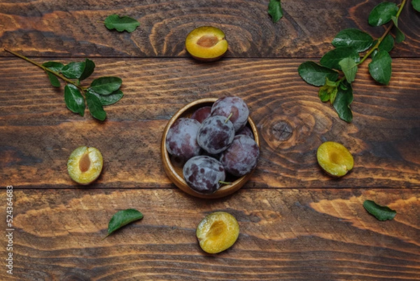 Fototapeta Ripe, juicy plums in bowl on brown wooden background with green leaves water drops, close-up, flatlay, rustic style, organic and veggie food concept, harvest season, ingredients for desserts, fructose