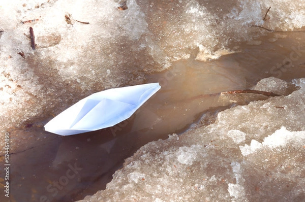 Fototapeta A white paper boat sails along a spring stream among the snowy shores