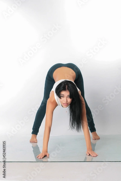 Fototapeta a pretty girl brunette stretching instructor, yoga, trainer, danser, posing in the studio on a white background in lasins and T-shirt