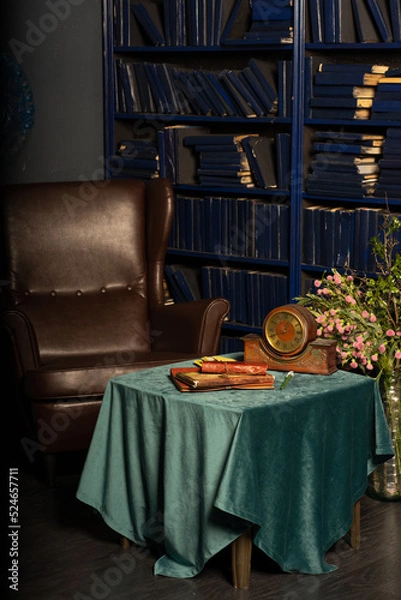 Fototapeta An old vintage gothic library with blue books on the shelves, a table and a brown leather armchair. On the table there is an old clock and vintage books in brown leather covers.