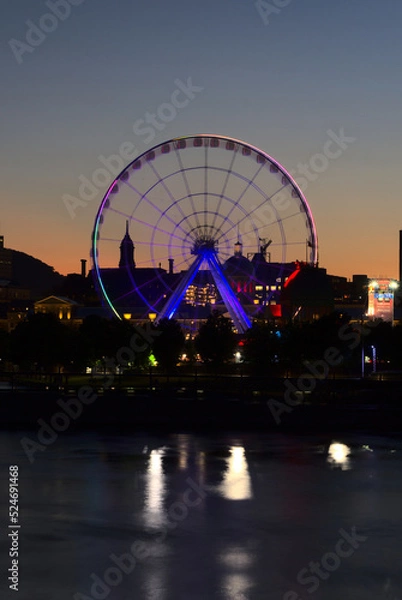 Fototapeta Long exposure of Montreal Grand Ferris Wheel in Old Port with motion blurat twilight blue hour. Reflections of sun rays on Saint Laurent river with a background of cloudy blue sky