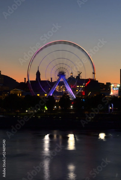Fototapeta Long exposure of Montreal Grand Ferris Wheel in Old Port with motion blurat twilight blue hour. Reflections of sun rays on Saint Laurent river with a background of cloudy blue sky