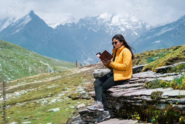 Obraz Portrait of Indian teenager girl tourist sitting in front of snowy mountains and reading book. Girl in mountains. Hobby and travel lifestyle concept