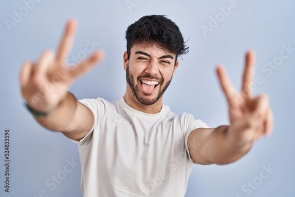 Fototapeta Hispanic man with beard standing over white background smiling with tongue out showing fingers of both hands doing victory sign. number two.