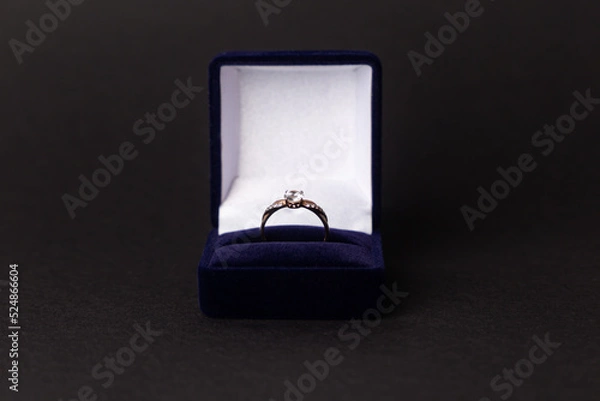 Fototapeta Jewelry, gold diamond ring in a blue box on a black background, isolated.  A ring for a marriage proposal. Close-up. Front view. A place for your text.