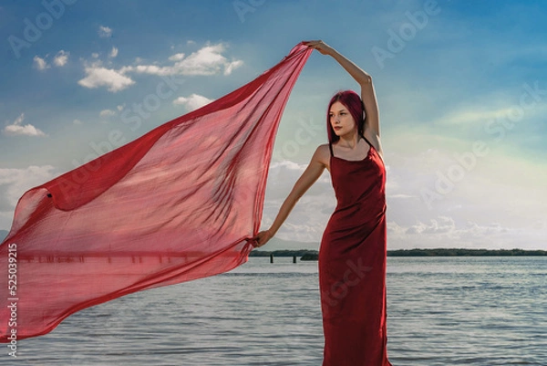 Fototapeta A beautiful graceful slender model girl in a silk or satin red dress with red hair poses and waves a red pareo handkerchief in the wind, against the background of the river and the sunset sky clouds