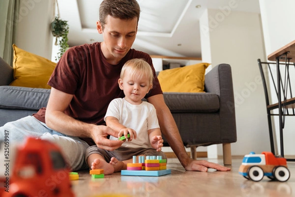 Obraz Father assisting his baby boy playing sorter sitting on floor in living-room with sofa on background, surrounded with car toys, waiting mom to come home from work. Education toys, child development