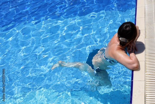 Fototapeta Woman in swimsuit relax in transparent water leaned on edge of swimming pool, top view. Girl swimmer in summer, beach vacation and cooling in hot weather