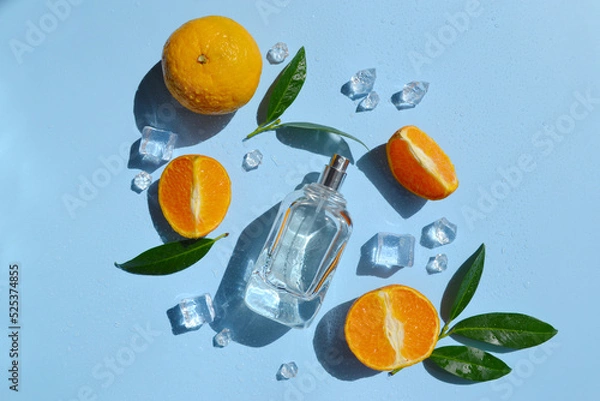 Fototapeta Glass bottle of perfume with oranges, green leaves, ice cubes, water drops on the light blue background with day sunlight. Smell of summer. Fresh orange cooling perfumery.  Direct sun rays. 