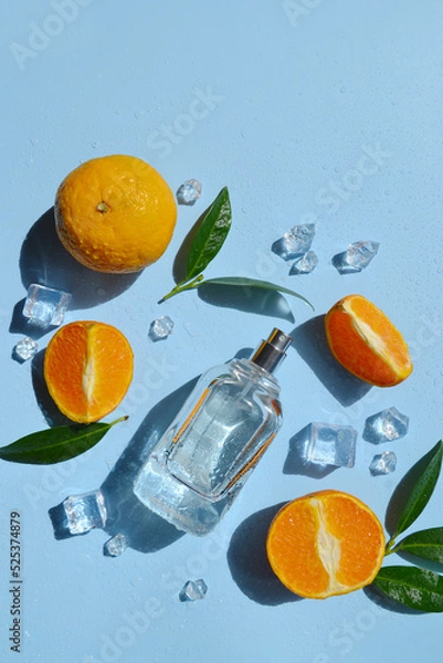 Fototapeta Glass bottle of perfume with oranges, green leaves, ice cubes, water drops on the light blue background with day sunlight. Smell of summer. Fresh orange cooling perfumery.  Direct sun rays. 