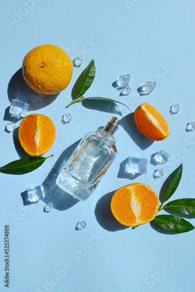 Fototapeta Glass bottle of perfume with oranges, green leaves, ice cubes, water drops on the light blue background with day sunlight. Smell of summer. Fresh orange cooling perfumery.  Direct sun rays. 