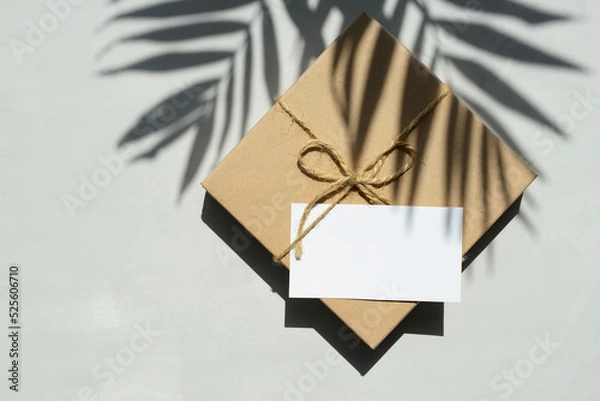 Fototapeta Gift box wrapped in kraft paper with blank greeting card and palm tree leaves shadows on a gray background