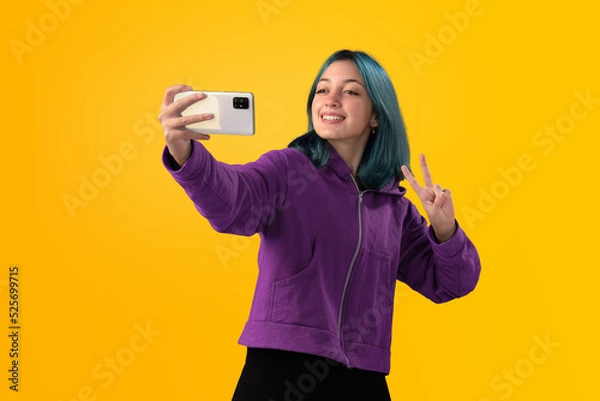 Fototapeta Cheerful nice young girl with blue hair taking selfie on cellphone and smiling. Isolated on yellow baclground