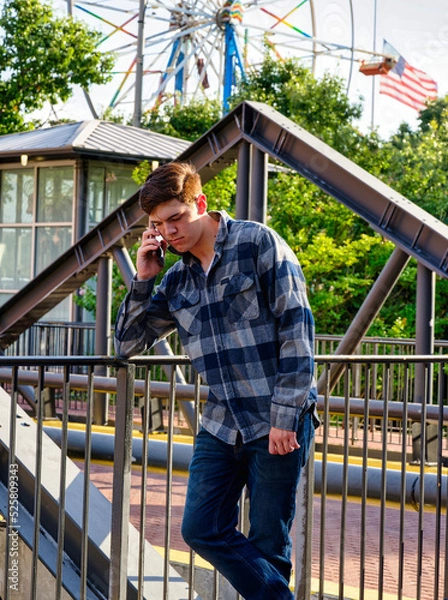 Fototapeta Handsome young man standing in front of iron railing and talking on a cellphone with a ferris wheel in the background.