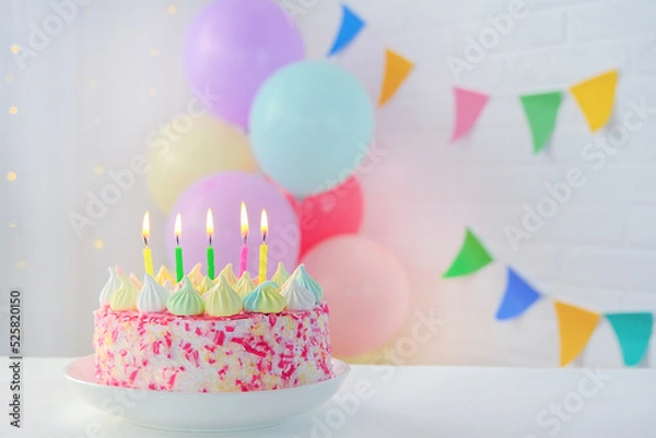 Fototapeta A bright, colorful cake with five lit candles sits on a table against a beautifully decorated wall with balloons and colored flags.