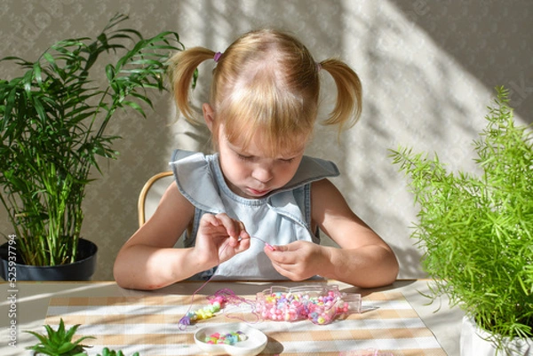 Fototapeta A two-year-old girl is sitting collecting beads from small beads. Development of fine motor skills in a child. DIY