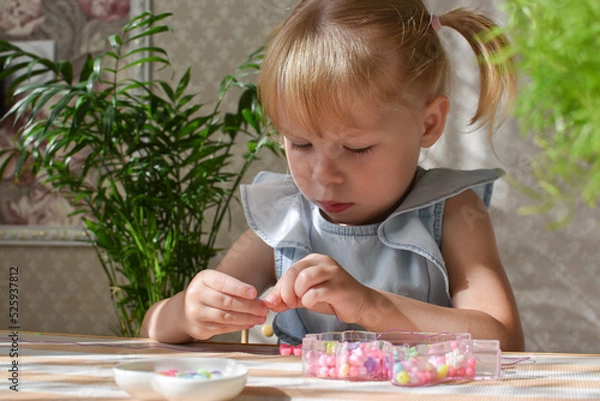 Fototapeta A two-year-old girl is sitting collecting beads from small beads. Development of fine motor skills in a child. DIY
