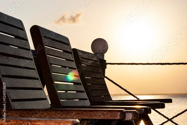 Fototapeta Holzliegen an der Küste während eines Sonnenuntergangs. Der Himmel ist gelb-orange und die Sonne groß und weiß. Die hölzernen Stühle nebeneinander am Strand oder Balkon und sind seitlich abgebildet.