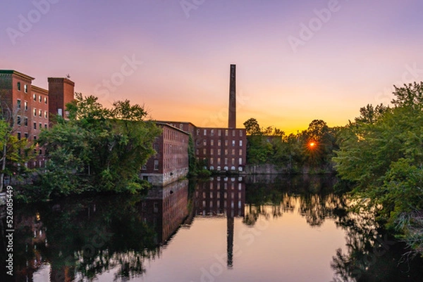Fototapeta A complex of historic cotton mill buildings with a chimney in an old industrial park with a reflection in the Nashua River late at sunset. Nashua, New Hampshire, USA