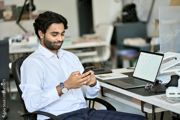 Fototapeta Indian young business man company worker, professional employee, stock market trader holding smartphone using cell phone mobile apps reading news, checking financial market data at work in office.