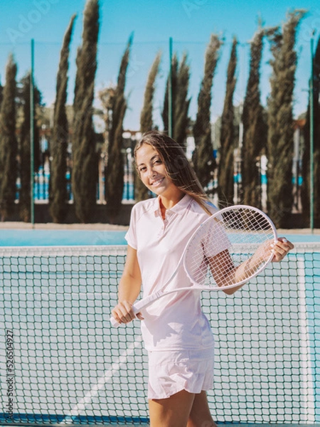 Fototapeta Mujer joven sonriente usando una raqueta de tenis en una pista de tenis al aire libre durante las vacaciones de verano