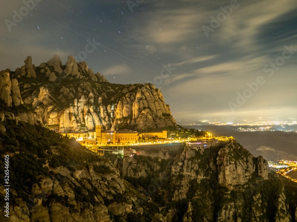 Fototapeta Paisaje nocturno de la montaña de Montserrat con fondo iluminado de poblaciones y cruz en primer termino en Cataluña (España).