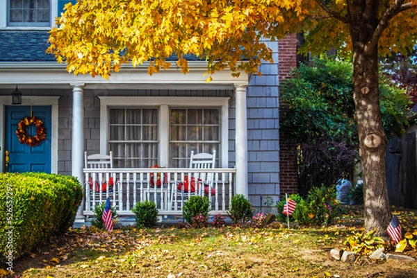 Fototapeta Blue shinngled two stor house entrance with porch and rocking chairs and American flags and a fall wreath under a yellow-leafed Autumn maple tree - closeup