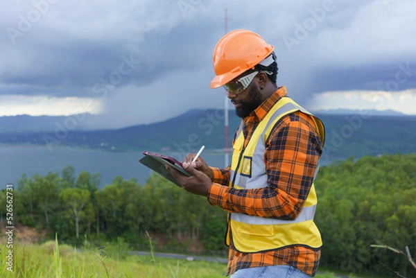 Fototapeta Black Engineer wearing safety jacket and hardhat holding tablet while working at outdoor, Smart working lifestyle.