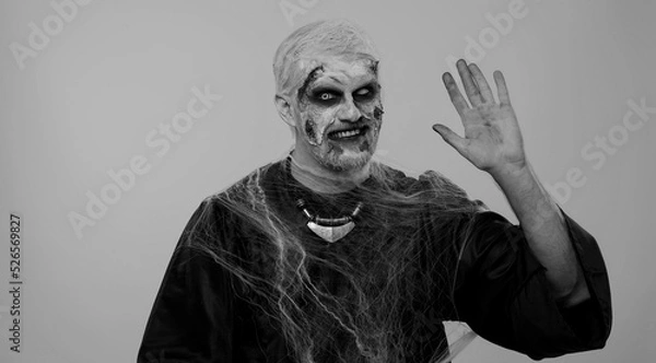 Fototapeta Creepy man with bloody scars face, Halloween zombie make-up. Scary wounded undead guy smiling friendly at camera and waving hands gesturing hello or goodbye, welcoming with hospitable expression